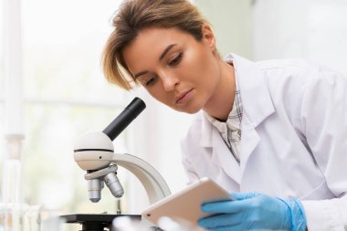 Kadın bilim adamı araştırma sırasında bir laboratuvarda mikroskop ve tablet bilgisayar kullanıyor.