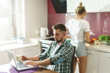 Sabahları mutfakta genç ve mutlu bir çift. Karısı lezzetli kahvaltı hazırlarken laptopunu kullanan yakışıklı bir adam..
