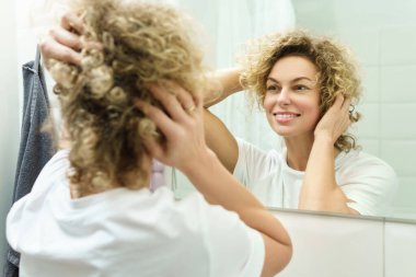Genç ve güzel bir kadın. Kıvırcık saçlı. Banyoda aynaya bakıyor.
