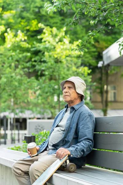 ベンチに腰掛けて街の公園でコーヒーを飲むロングボードを持つスタイリッシュな高齢者 — ストック写真