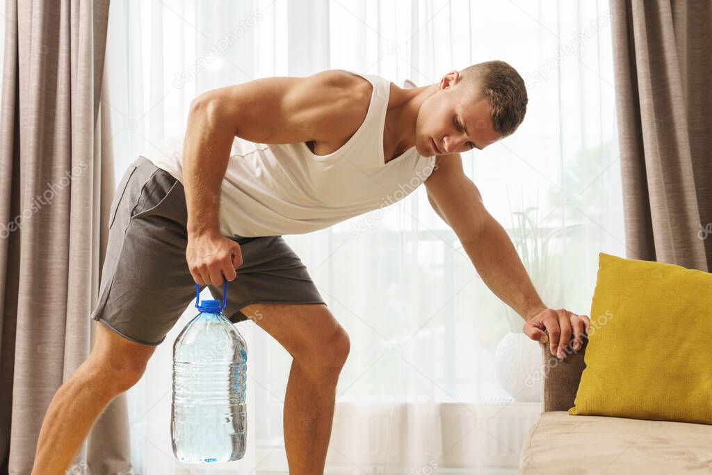 Young athletic man using big bottle of water like an alternative of dumbbell for home workout