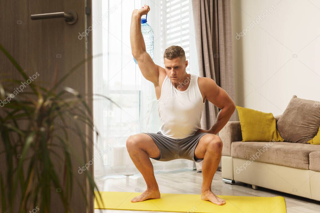 Young athletic man using big bottle of water like an alternative of dumbbell for home workout