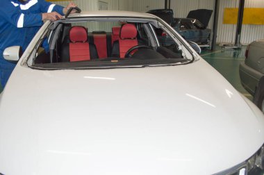 Mavi üniformalı tamirci araba tamirhanesinde beyaz bir arabanın ön camını sökmüş.