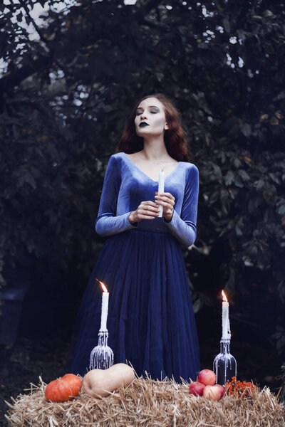 A girl in the image of a witch. The idea of a photo shoot for Halloween. Terribly beautiful. Blue dress, candles, pumpkins, smoke. Halloween attributes. Art photo session