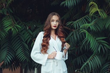 Büyük yeşil yaprakların arka planında beyaz önlüklü muhteşem bir kadın. Kupa ve siyah bardaklar ellerde. Kırmızı ruj ve oklar. Sabah fotoğrafı. 