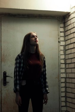 Genç bir kadın loş bir koridorda bir kapının yanında duruyor ve geceleri bir binanın tavanına bakıyor.