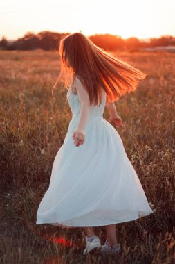 Pretty cute girl is circling in a white dress on the field. Sun rays. Orange sunset. Concept. A woman dances joyfully