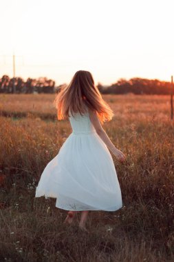 Pretty cute girl is circling in a white dress on the field. Sun rays. Orange sunset. Concept. A woman dances joyfully