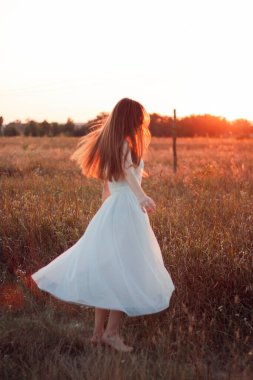 Pretty cute girl is circling in a white dress on the field. Sun rays. Orange sunset. Concept. A woman dances joyfully