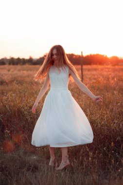 Pretty cute girl is circling in a white dress on the field. Sun rays. Orange sunset. Concept. A woman dances joyfully