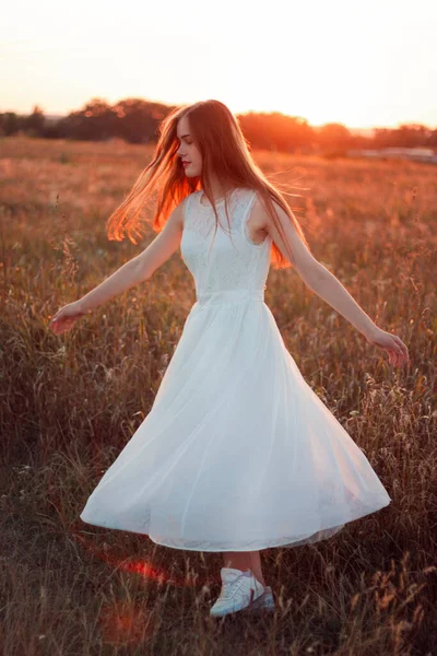 Pretty cute girl is circling in a white dress on the field. Sun rays. Orange sunset. Concept. A woman dances joyfully