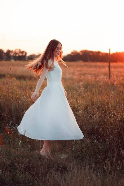 Pretty cute girl is circling in a white dress on the field. Sun rays. Orange sunset. Concept. A woman dances joyfully