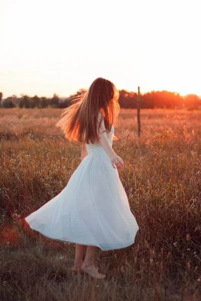 Pretty cute girl is circling in a white dress on the field. Sun rays. Orange sunset. Concept. A woman dances joyfully