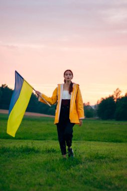 Ukraynalı kadın büyük bir Ukrayna bayrağıyla sahada. Vatansever fotoğraflar. Kahramanlık ve özgürlük. Kavram. Sarı yağmurluk giymiş güzel bir kadın. Seyahat
