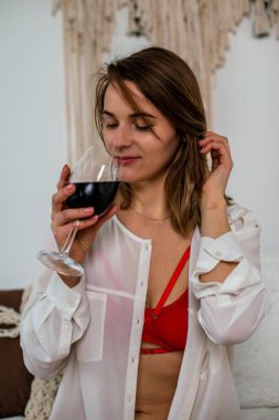 Girl in red lingerie and a white shirt with a glass of red wine sitting on the bed.