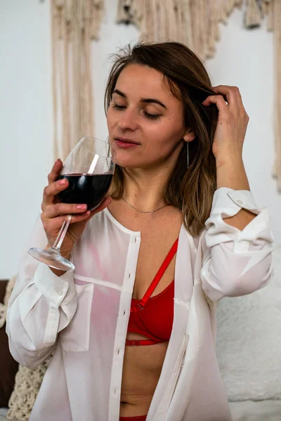 Girl in red lingerie and a white shirt with a glass of red wine sitting on the bed.