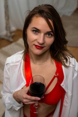 A girl in red underwear and a white shirt with a glass of red wine is sitting on the floor.