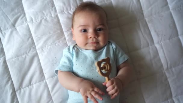 Bebê bonito recém-nascido de três meses de idade, com um chocalho natural de madeira, encontra-se na cama e olha para a câmera, close-up, vista superior — Vídeo de Stock