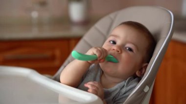 Elinde yeşil kaşık olan sevimli bebek mutfaktaki sandalyede oturur ve kameraya bakar.