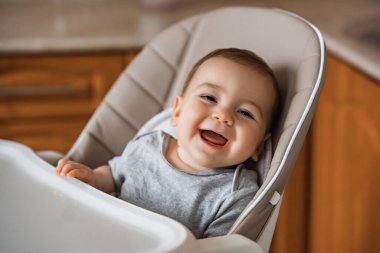 Mutfakta yüksek sandalyede oturan mutlu bir bebek. Çocuklar için sağlıklı beslenme. Çocuklar, çocukların güvenliği. Küçük çocuk oynuyor ve evde öğle yemeği yemeye hazır.