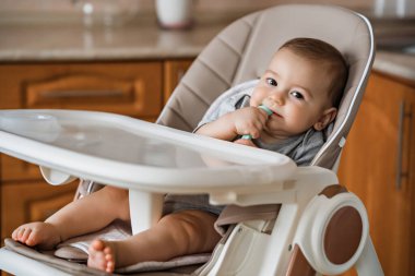 Elinde kaşık olan sevimli bir bebek yüksek bir sandalyede oturur ve kameraya bakar. Bebeklerin ilk beslenmesi konsepti