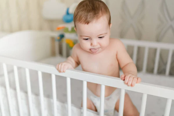 Pequeno bebê europeu em uma fralda fica em um berço em casa, bebê saindo da língua — Fotografia de Stock