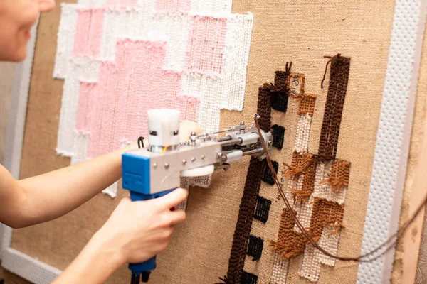 The process of making a tufting rug. Tufting a pistol in female hands against the background of a handmade brown tank rug on sacking.
