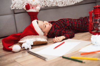 Noel Baba şapkalı ve kırmızı kareli pijamalı küçük bir çocuk Noel Baba 'ya mektup yazarken yerde uyuyakalmış..