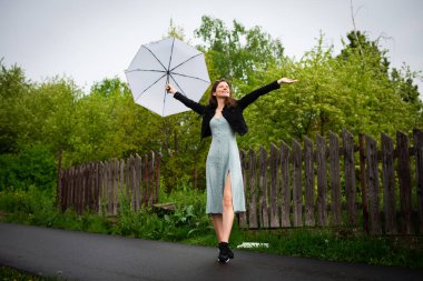 Hafif narin elbiseli mutlu bir genç kadın yağmurlu havada hafif bir şemsiyeyle yürür..