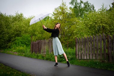 Hafif narin elbiseli mutlu bir genç kadın yağmurlu havada hafif bir şemsiyeyle yürür..