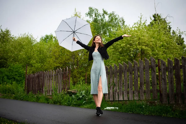 Hafif narin elbiseli mutlu bir genç kadın yağmurlu havada hafif bir şemsiyeyle yürür..