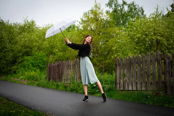 Hafif narin elbiseli mutlu bir genç kadın yağmurlu havada hafif bir şemsiyeyle yürür..