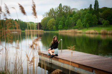 Dalgalı saçlı genç bir kadın romantik bir elbiseyle bir iskelede tek başına oturuyor..
