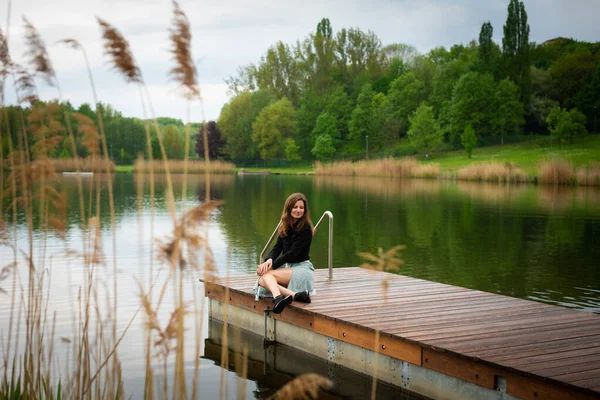 Dalgalı saçlı genç bir kadın romantik bir elbiseyle bir iskelede tek başına oturuyor..