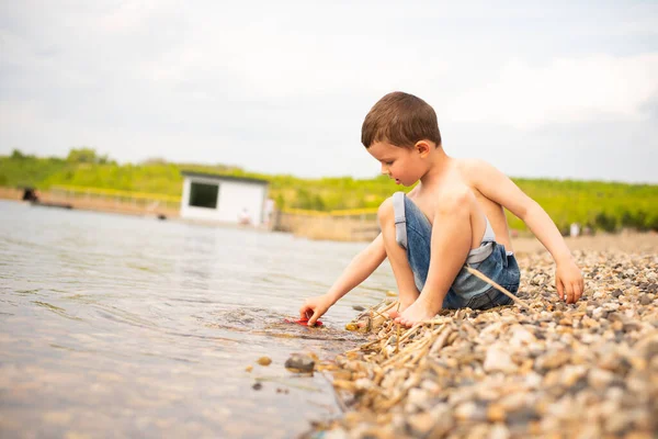 A small six-year-old boy in blue denim shorts sits on a stone seashore.