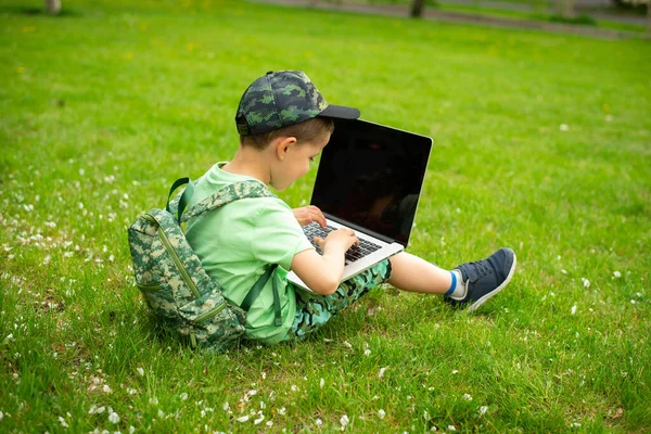 A cheerful kid with a laptop sits on green grass in green clothes and a cap.