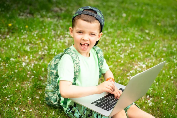 A cheerful kid with a laptop sits on green grass in green clothes and a cap.