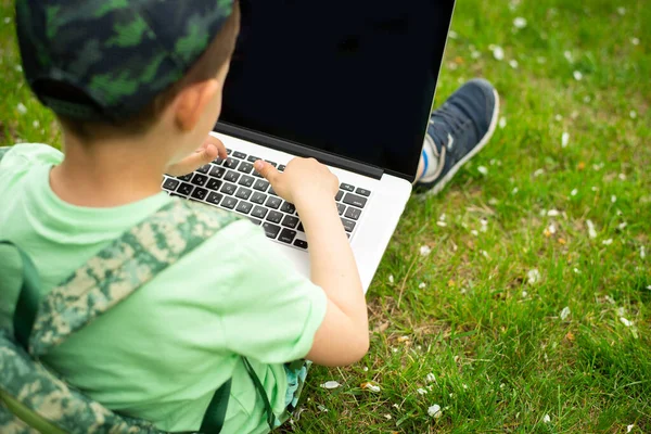 A cheerful kid with a laptop sits on green grass in green clothes and a cap.