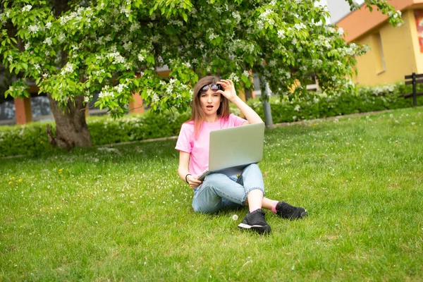 Surprised young woman works on a laptop while sitting on green grass in jeans and a pink T-shirt against the backdrop of a flowering tree.