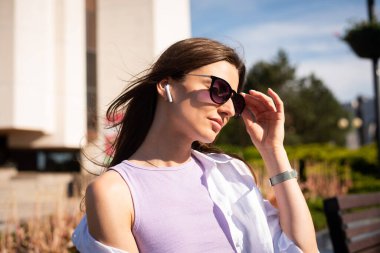 Şehir meydanında güneşli bir havada, kablosuz kulaklıklar ve güneş gözlükleriyle genç, zayıf bir kadın bankta oturuyor..