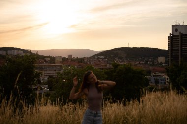 Gün batımında şehrin arka planında poz veren neşeli genç kadın dağlarda.