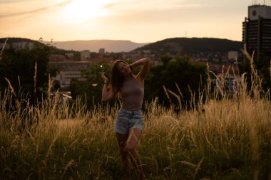 Gün batımında şehrin arka planında poz veren neşeli genç kadın dağlarda.