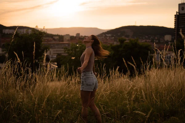 Genç, neşeli bir kadın gün batımında, dağlardaki bir şehrin arka planında dönüyor..