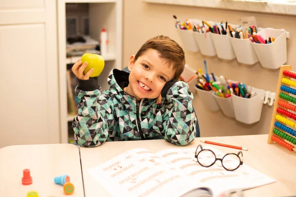 Little Schoolboy Sits His Desk Smiles Holding Apple Snack His — Stockfoto