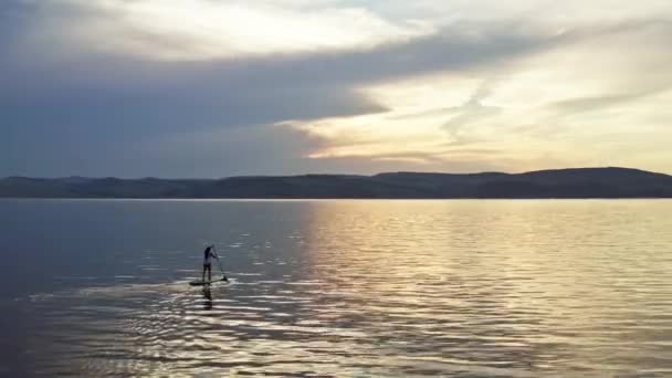 Drone disparo de una silueta de una joven mujer flotando en un tablero con una pala en un lago de montaña al atardecer. — Vídeos de Stock