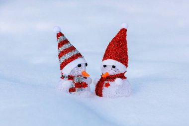 Noel arkaplanı, kırmızı şapkalı iki kardan adam ve kar yığınında bir eşarp, kar üzerinde Noel Baba gibi giyinmiş kardan adamlar.