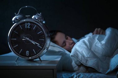 Genç bir kadın gece yatağında ay ışığında uyuyor, ön plandaki çalar saati kapatıyor.