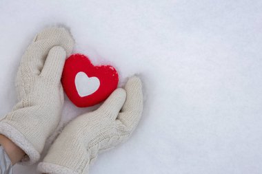 hands in warm knitted mittens hold a red heart on a snow background, concept of love and valentines day background