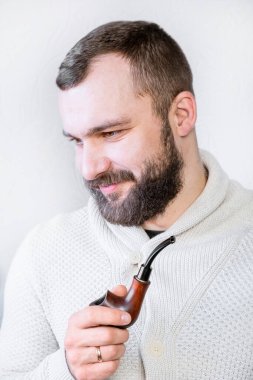 Hafif hırkalı, sakallı ve bıyıklı gülümseyen bir beyefendi pipo tutuyor ve sigara içiyor, şık, modaya uygun bir adam.