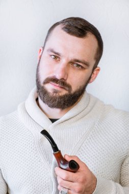 Hafif hırkalı, sakallı ve bıyıklı gülümseyen bir beyefendi pipo tutuyor ve sigara içiyor, şık, modaya uygun bir adam.